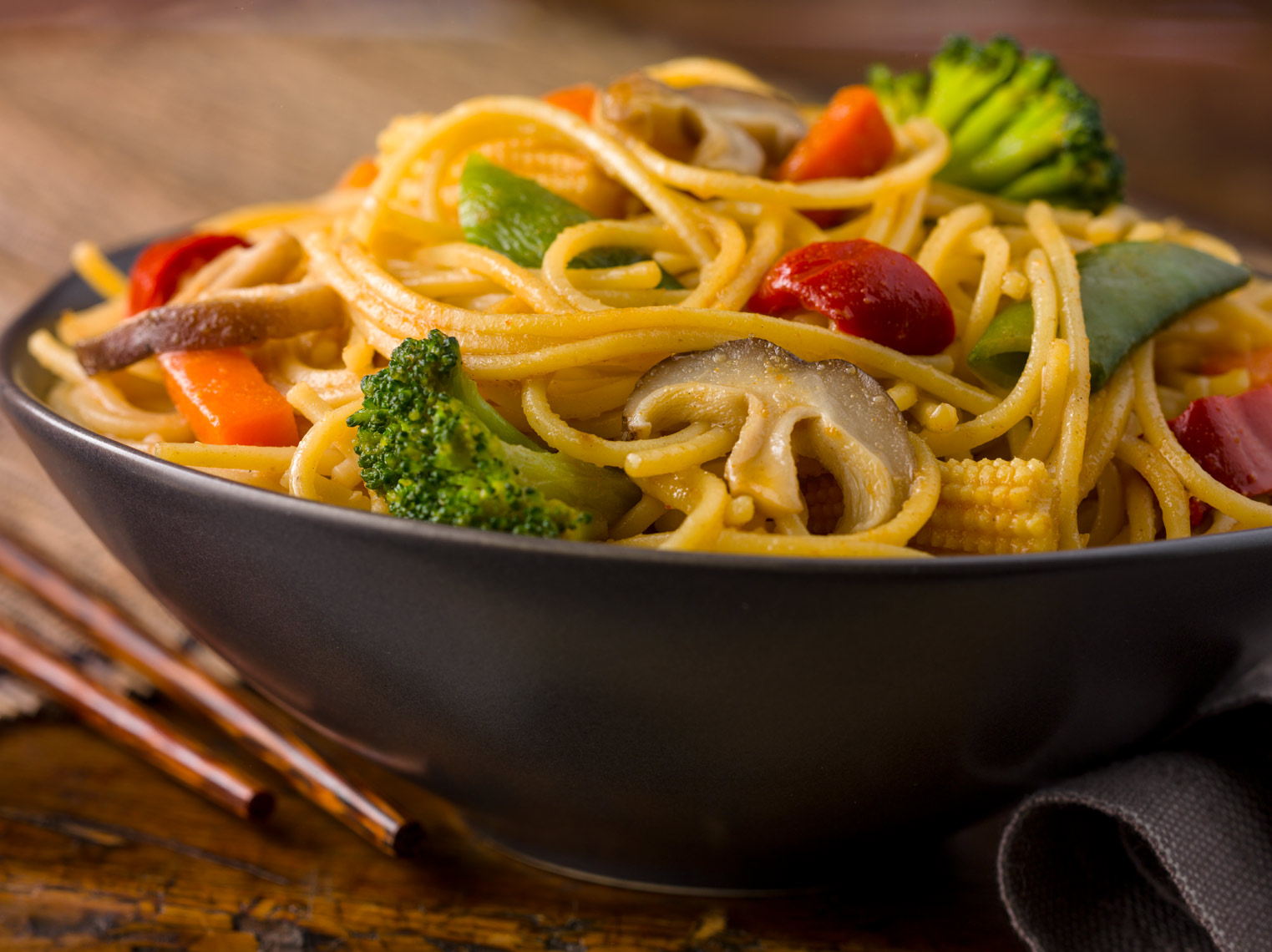 vegetable lo mein with red peppers, carrots, mushrooms, snow peas, baby corn, and broccoli in black bowl on wood surface with chopsticks Fantich studio food photography.